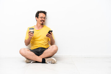 Young caucasian man sitting on the floor isolated on white background holding coffee to take away and a mobile