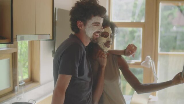 Medium shot of young multiethnic couple with clay masks on faces posing for smartphone camera while taking selfie in kitchen at home