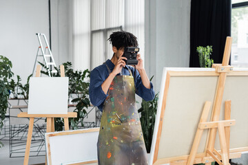 Young african american artist in apron taking photo on vintage camera near canvas in studio.