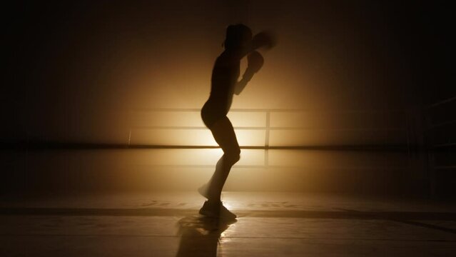 Silhouette of a girl having intensive workout in gloves. Close-up shot of strong, aggressive woman training endurance and power of her body muscles. High quality 4k footage in golden foggy back light
