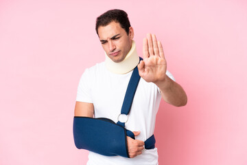 Young caucasian man with broken arm and wearing a sling isolated on pink background making stop...