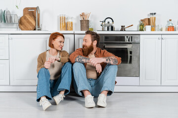 Smiling adult couple looking at each other while sitting on floor in kitchen.