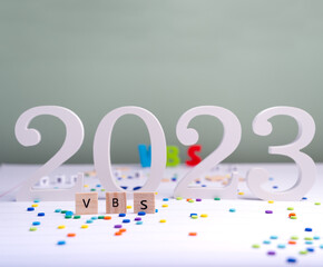 Wooden cube with text VBS (Vacation Bible School) 2023 and confetti on a white table. Christian concept.