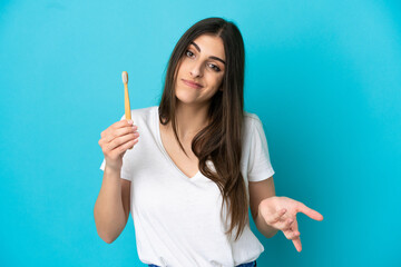 Young caucasian woman brushing teeth isolated on blue background making doubts gesture while lifting the shoulders