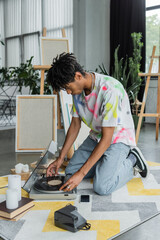 African american artist using record player near books and canvases in workshop.