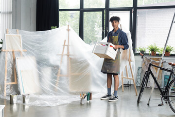 Young african american artist in apron holding box near paints and cellophane in studio.