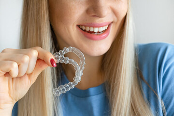Cropped photo of young beautiful woman smiling with hand holding dental aligner retainer (invisible) on white background of dental clinic for beautiful teeth treatment course concept - obrazy, fototapety, plakaty