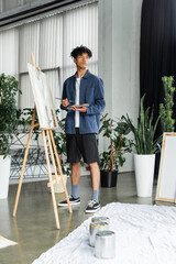 Young african american artist holding paintbrush and palette near canvas on easel in studio.