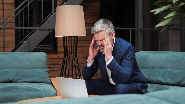 Serious tired businessman office worker sitting at workplace working on laptop massages forehead having severe pain. Stressed business man or freelancer solving difficult problem, feeling headache.