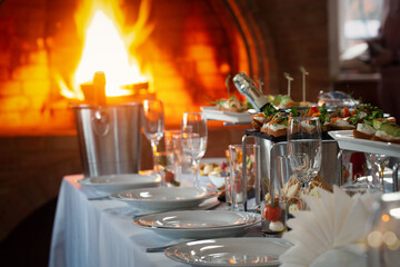 Set banquet table with plates, wine glasses, appetisers, cutlery and a buckets of champagne indoor next to the fireplace, selective focus.