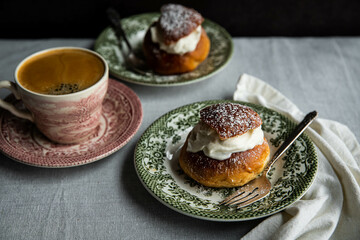 Homemade semlor buns with frangipane and whipped cream on vintage plate and cup of coffee on linen tablecloth.