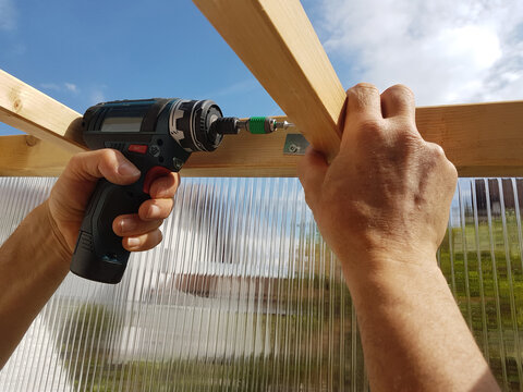 man builds a greenhouse out of wooden beams and double-walled panels. fixing the screws with a drill