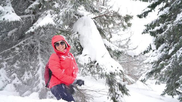 Smiling young woman in sunglasses dressed warm winter trekking red jacket beating a spruce branch covered with snow starting snowfall. Happy wintertime vacation concept 4K footage.
