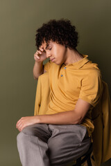 brunette african american man in yellow polo shirt sitting with closed eyes and hand near face on grey green background