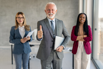 Coworkers smiling cheerfully in office. Happy senior businessman holding laptop and showing thumb up while standing in a modern workplace with his female colleagues. Businesspeople working as a team.