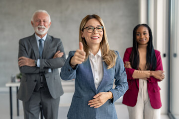 Fototapeta na wymiar Shot of beautiful confident smiling businesswoman wearing glasses in suit standing in front of her successful team showing thumb as sign of success.