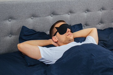 A man wrapped in a blanket wearing a sleep mask on a gray background. Sleeping at home in his bed. The concept of rest and relaxation.