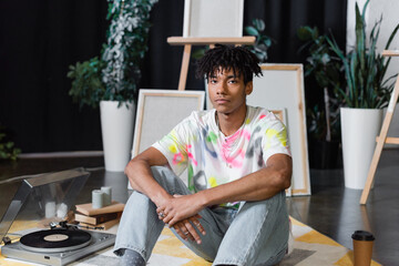 African american artist looking at camera near vinyl player and books in studio.