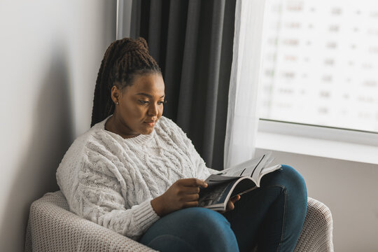 African American Woman With Dreadlocks Reading Magazine At Home - Leisure And Lifestyle Concept