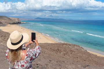 Fototapeta na wymiar Turista rubia con pelo largo, vestido de flores y sombrero de paja tomando una foto con su teléfono móvil de la playa de arena blanca de Jandia con mar turquesa en Fuerteventura, Islas Canarias.