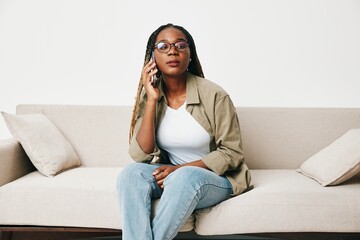 Obraz premium African American woman business freelancer working sitting on the couch at home in the phone, business calls and messages happiness smile, home clothes and eyeglasses, light interior background.