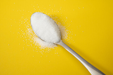 A silver stainless steel spoon filled with sugar or salt that is overflowing on to a yellow...