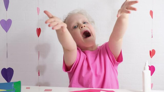 cute girl in a pink t-shirt shows a heart with her palms sitting at the table, preparing for valentine's day