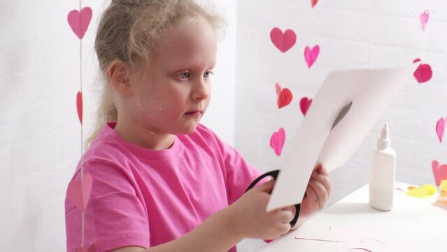 girl with blond curly hair cuts something out of cardboard, decor for valentine's day, decorated room