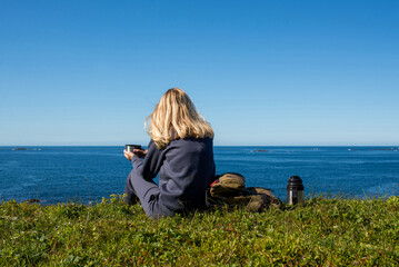 Woman with a cup of hot tea and a thermos sits on the ocean and admires the picturesque landscape. Beautiful nature. Harmony, relaxed lifestyle. Travel, adventure. Explore Northern Norway. Scandinavia