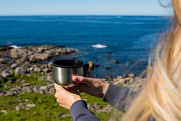 Woman with a cup of hot tea and a thermos sits on the ocean and admires the picturesque landscape. Beautiful nature. Harmony, relaxed lifestyle. Travel, adventure. Explore Northern Norway. Scandinavia