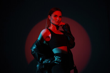 Fototapeta na wymiar Fashionable beautiful creative woman in trendy black clothes with stylish top and leather jacket posing in studio with color red and blue light in studio