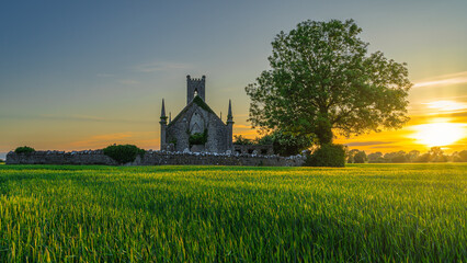 Cereal field illuminated by sunlight and old stone ruins of Ballinafagh Church with dramatic sky at sunset in background, County Kildare, Ireland - obrazy, fototapety, plakaty