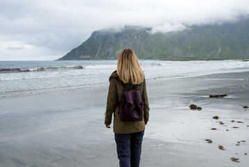 Blond hair girl with a backpack walks along an empty sandy ocean beach. Town and mountains. Beautiful nature landscape. Lonely girl. Wanderlust. Travel, adventure, lifestyle. Explore North, Norway 