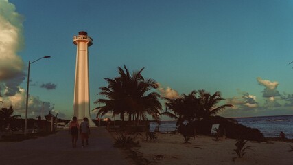 Fotografia de un faro con la mar y precencia de palmeras marcadas por las sombras al costado derecho