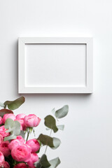 White photo frame mockup hanging on white wall and and bouquet of pink roses with eucalyptus leaves. Empty blank frame and flowers