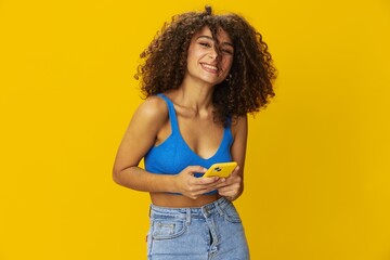 Influencer woman with phone in hand taking selfies talking on video call freelancer social media technology work, with curly afro hair in blue shirt yellow background , smile with teeth, copy space