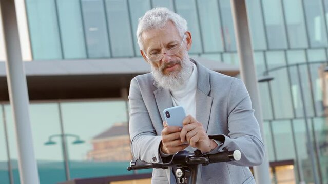 Old senior business man holding smartphone using electric bike rental digital phone mobile app renting scooter in city hiring public eco transport mobile application standing in business district.