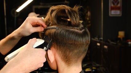 men's hairstyling and haircutting with hair clipper in a barber shop or hair salon. Hairdresser service in a modern barbershop in a dark key lightning with warm light back view
