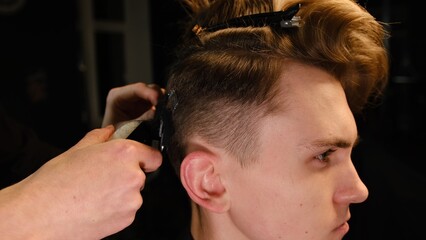 Fototapeta premium men's hairstyling and haircutting with hair clipper in a barber shop or hair salon. Hairdresser service in a modern barbershop in a dark key lightning with warm light side view