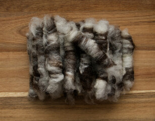 A stack of small brown and white rolags made of sheep's wool, ready for spinning, against a wooden background.
