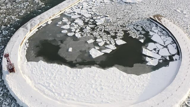 Panning up aerial shot of a hooked or curved pier covered in ice and snow in Lake Michigan with angular and rounded chunks of ice floating on the surface.