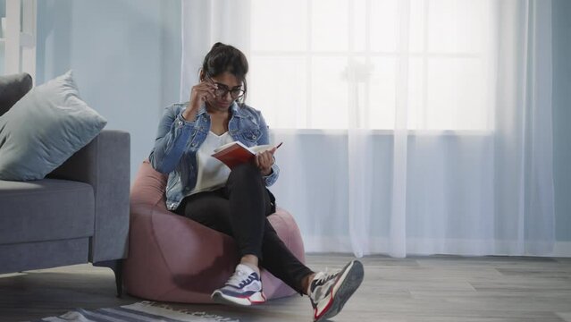 Relaxed Indian woman reads book sitting on bean bag chair in living room against bright window. Young lady reader enjoys captivating tale at home