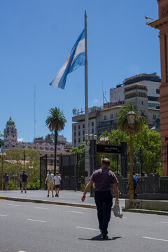 Hombre atravensando la calle, de fondo personas caminando, la plaza de mayo en buenos aires, y la bandera de argentina.