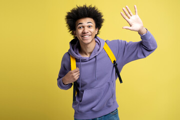 Smiling friendly African American man wearing stylish hoodie, waving hand isolated on yellow...