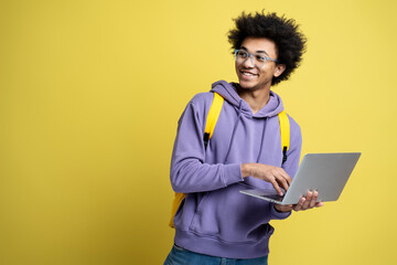 Handsome smiling African American man holding laptop computer working online looking away, copy space. Happy confident university student studying isolated on yellow background. Online education