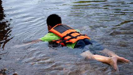 ฺBoy asia lying face down in the water wearing an orange life jacket. water on the banks of the...