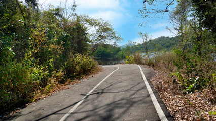 Path of a small asphalt road with forward bends. On both sides of the road there are trees and green grass. Background is a river and an island under the blue sky.