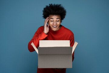 Young happy African American man influencer unpacking delivery box isolated on blue background. Excited customer opening package with birthday gift. Online shopping, sale concept   