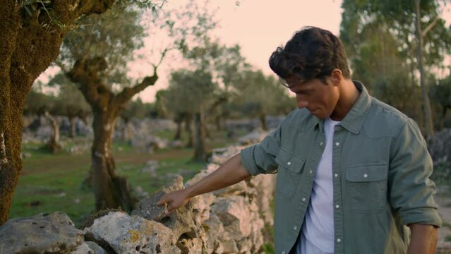 Thoughtful guy touching stone wall at garden portrait. Relaxed man walking alone