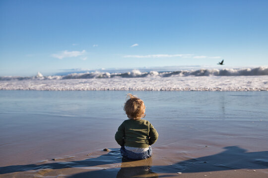 Blonde Kid Sitting on Sandy Beach and Watching the Waves